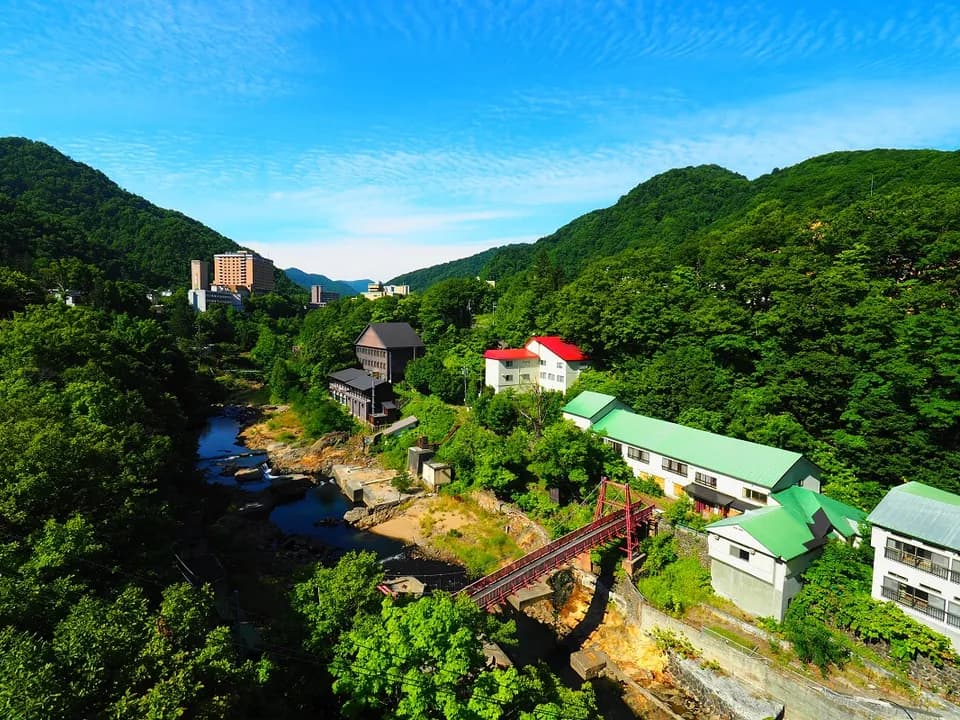 自定山溪大桥俯瞰的夏日溪谷。蓝天与青山之间流淌着丰平川，可见温泉街的建筑与红色桥梁
