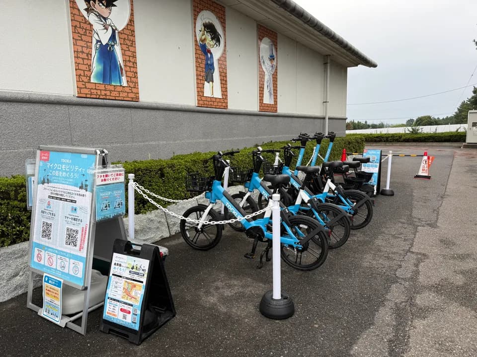 青山剛昌ふるさと館①のポート。キャラクターの壁画が並ぶ館の外壁の前に TOCKLE が6台並び、右奥に立入禁止のバーとコーン、左手に案内看板が置かれている