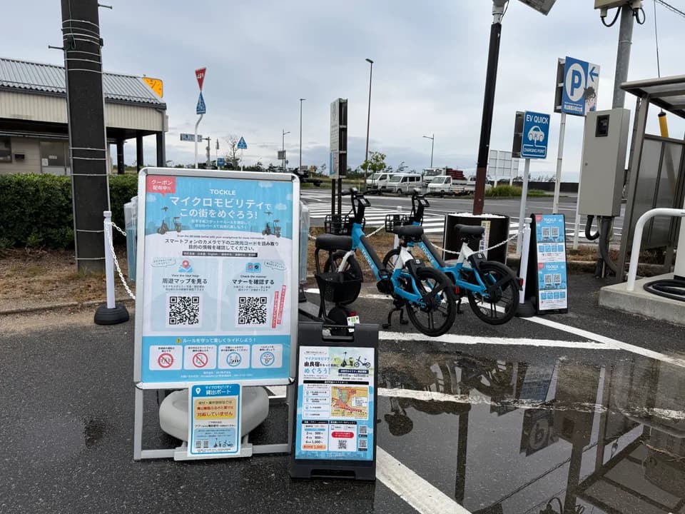 青山剛昌ふるさと館②のポート。雨上がりの駐車場で TOCKLE が2台、丸いごみ箱の脇に停まり、奥に交差点とEV充電スタンド、案内看板が並ぶ