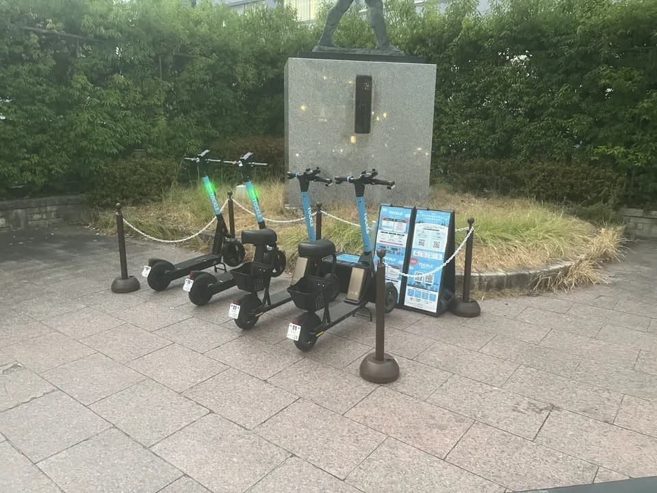 さが維新広場のポート。石畳の広場に TOCKLE が4台並び、奥に銅像を載せた御影石の台座と生垣が続く