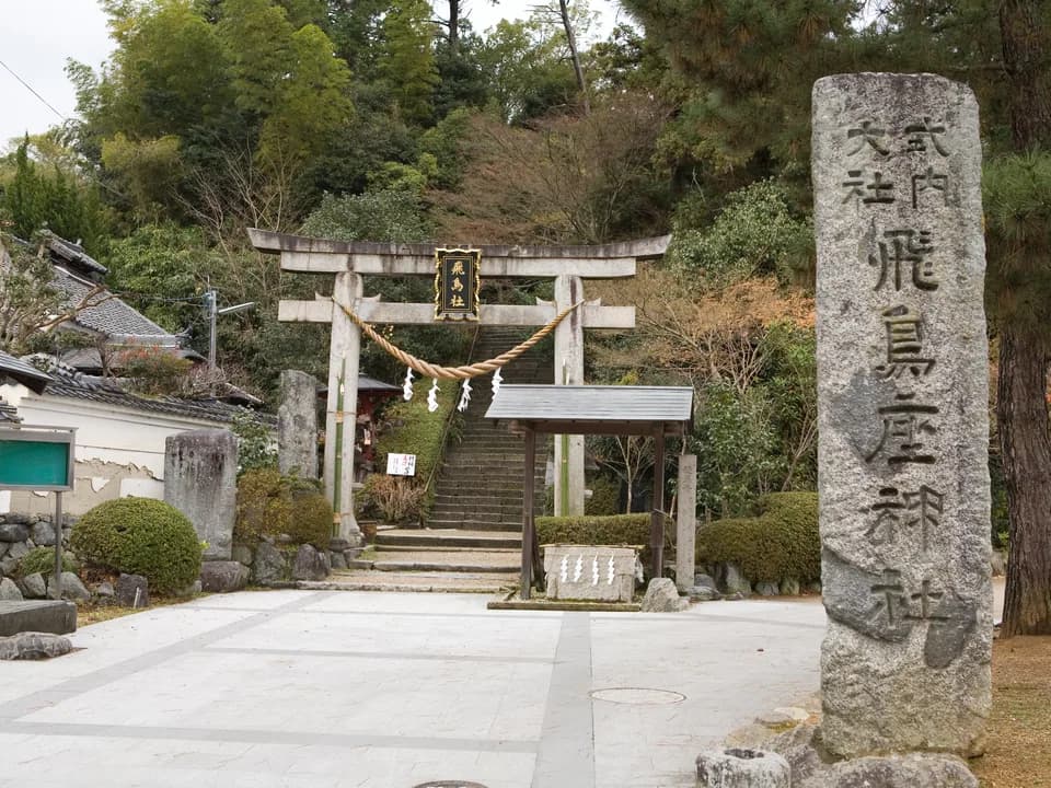 飛鳥坐神社の入口。注連縄を張った石の鳥居の奥に石段が続き、右手の大きな石柱には「飛鳥坐神社」と刻まれている。周囲は木々の緑に囲まれる
