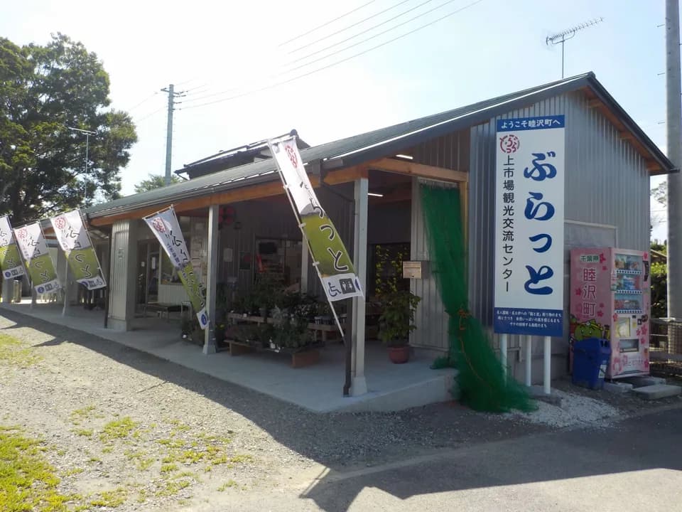 上市場観光交流センターぶらっとの外観。「ぶらっと」と大書きされた青い看板が建物の脇に立ち、のぼり旗が並ぶ軒先に鉢植えと苗が置かれている