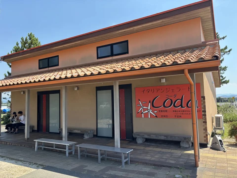 オレンジ色の瓦屋根と土壁の平屋。軒下にベンチが並び、右手の壁に風車の絵と「イタリアンジェラート Coda」と書かれた赤い看板が掛かる