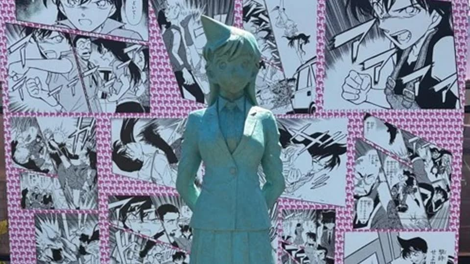 マンガのコマを貼りつめた壁を背に立つ、制服姿の少女のブロンズ像。緑青色の像が日差しを受けている