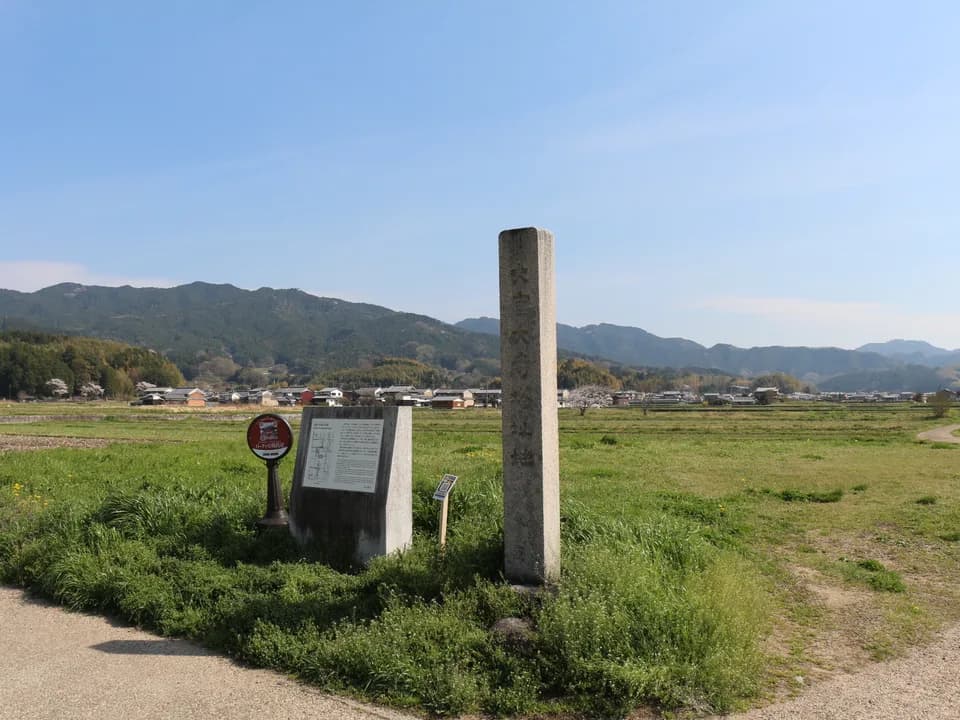大官大寺跡の草地に立つ石の標柱と説明板。奥には田畑と集落、青い山並みが広がり、空は晴れている
