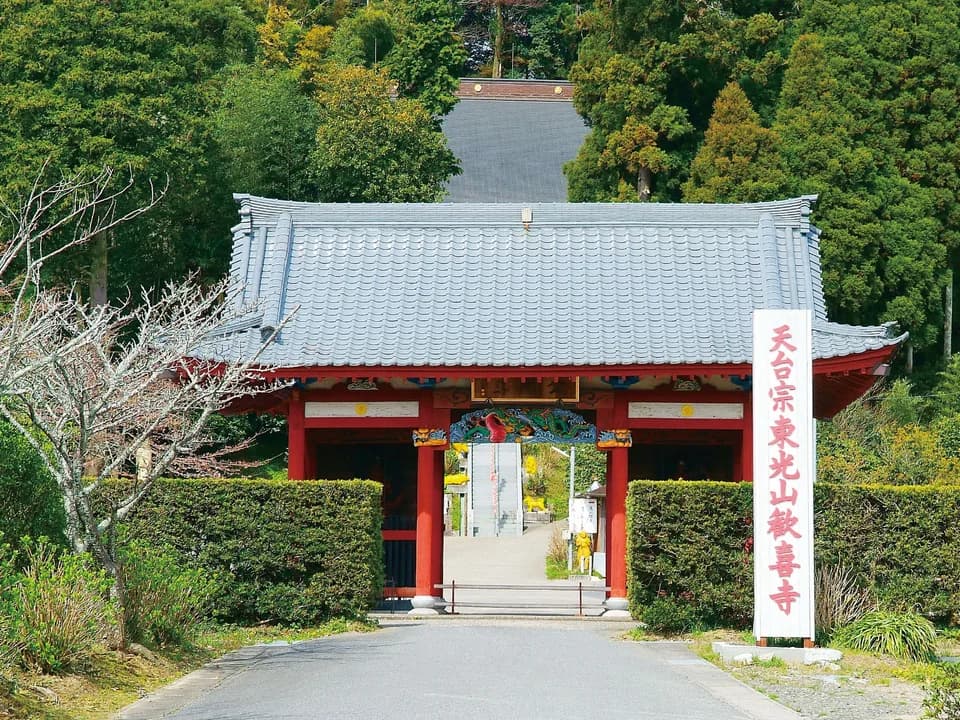 歓喜寺の仁王門。朱塗りの柱と灰色の瓦屋根、梁には彩色された龍の彫刻。右手に「天台宗東光山歓喜寺」と縦書きされた白い標柱が立ち、門の奥に石段が続く