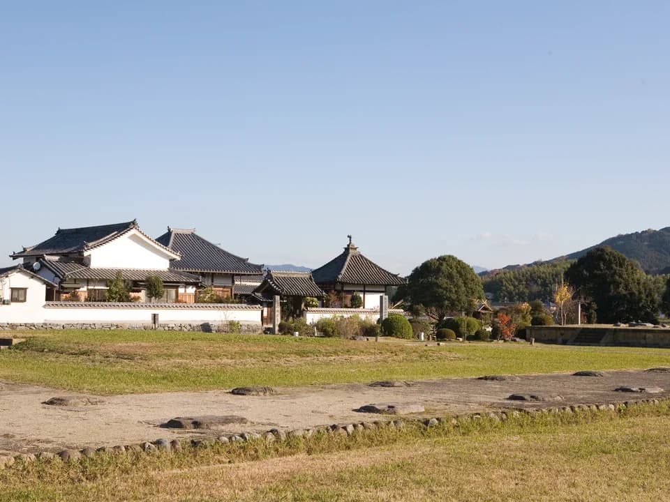 川原寺跡の景色。芝地に礎石が点々と並び、奥には白壁と瓦屋根の民家や小さな堂、木々、山並みが見える