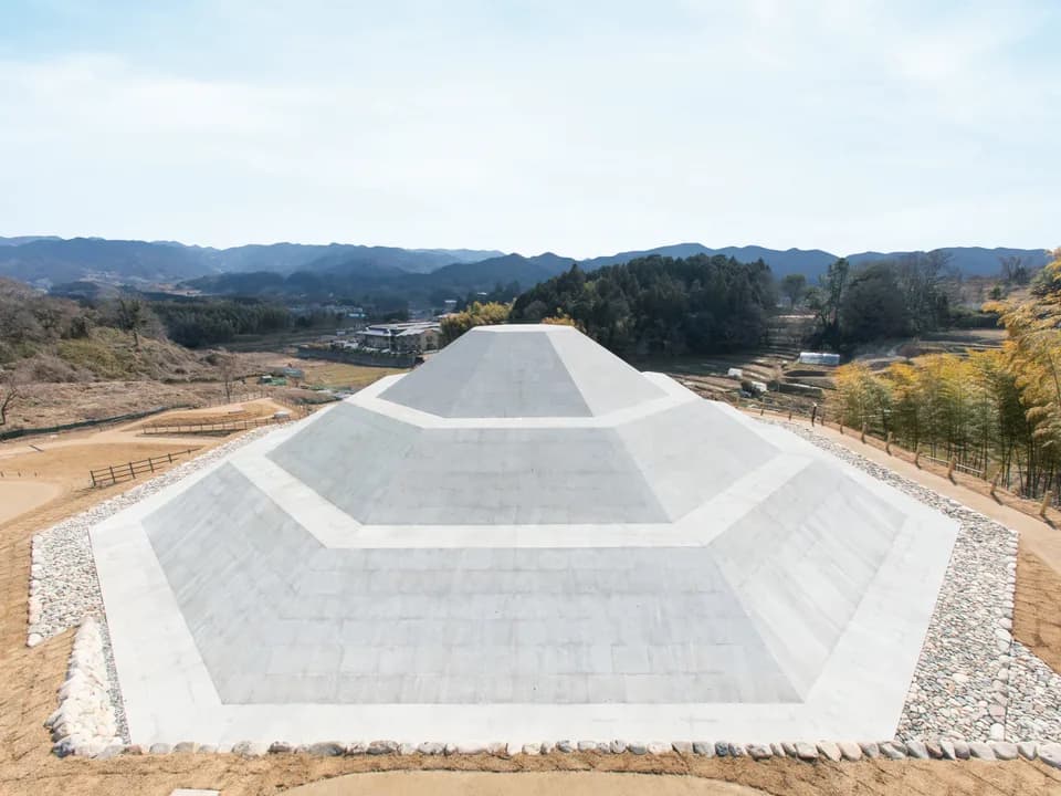 牽牛子塚古墳を高い位置から見たところ。白い石張りの八角形の墳丘が段状に立ち上がり、周囲を小石が縁取る。奥には山並みが続く