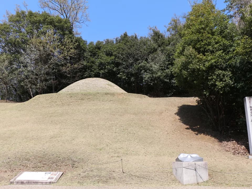 キトラ古墳の墳丘を正面から見たところ。枯れ色の芝に覆われた小さな丸い墳丘が木立を背に立ち、手前に説明板と石の台座、右端に石の標柱が見える