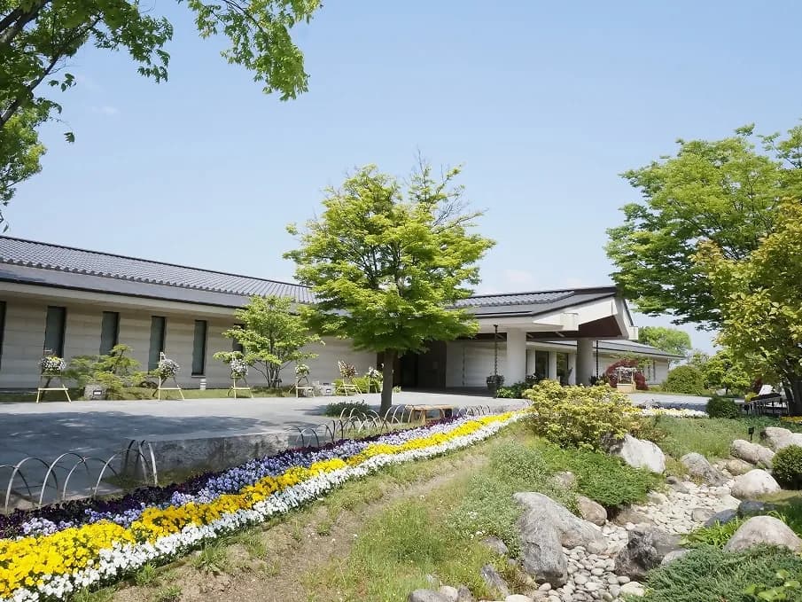 奈良県立万葉文化館の外観。灰色の瓦屋根をのせた低い建物の前に、黄・白・紫のパンジーの花壇が広がり、若葉の木々と石組みの庭が建物を囲む。空は青く晴れている