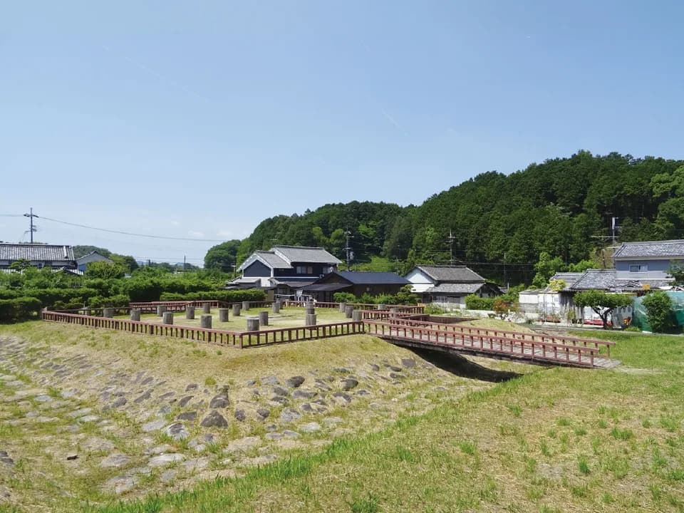 飛鳥水落遺跡の全景。赤茶色の柵で囲まれた区画に木の柱が並び、手前には石積み、奥には民家と緑の山が見える