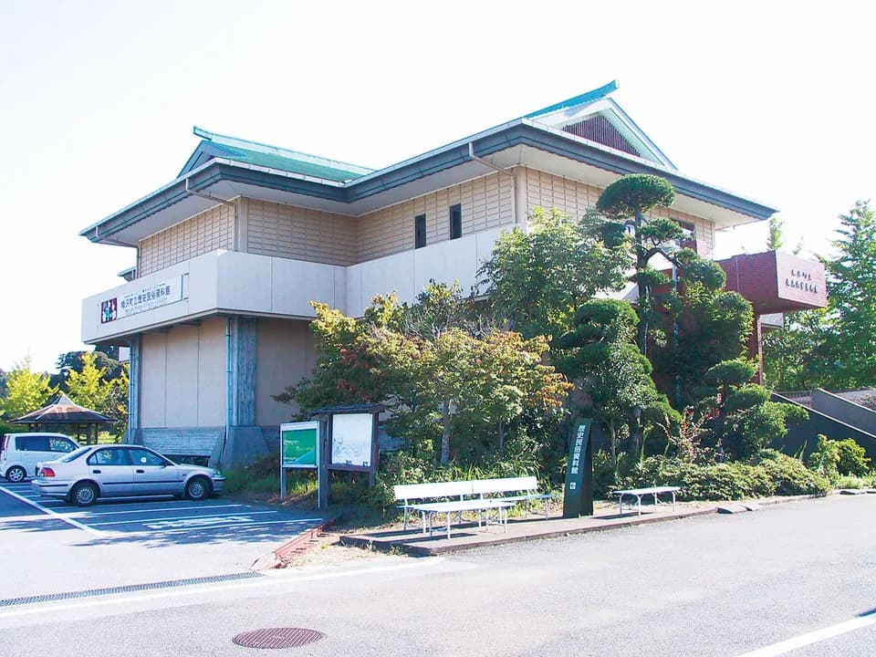 睦沢町立歴史民俗資料館の外観。緑青色の瓦屋根を重ねた2階建てで、入口脇に案内板とベンチ、手前は駐車場