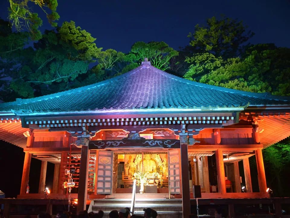 ライトアップされた妙楽寺の本堂。屋根が青緑、柱が朱色に照らされ、開いた扉の奥に金色の大日如来坐像が浮かび上がる。手前に見物する人影が並ぶ