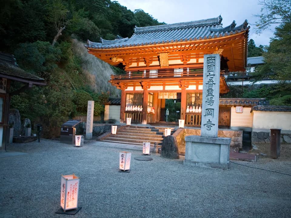 岡寺の山門を正面から見たところ。朱色の二層の門が夕暮れの光に浮かび、手前に「西国七番霊場 岡寺」と刻まれた石柱と、灯りのともる行燈が並ぶ