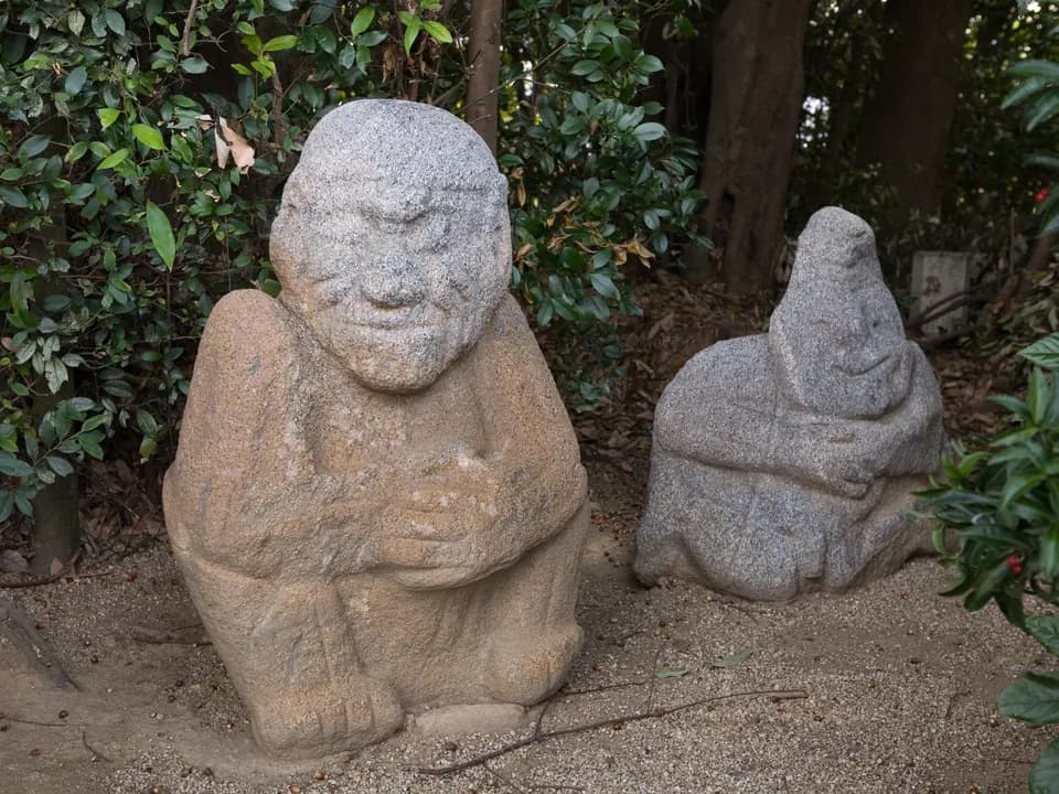 吉備姫王墓にある猿石。笑みを浮かべた顔と、腕を前で組んで座る姿の石像が2体、砂利の地面に並ぶ。背後は常緑の生け垣と木立