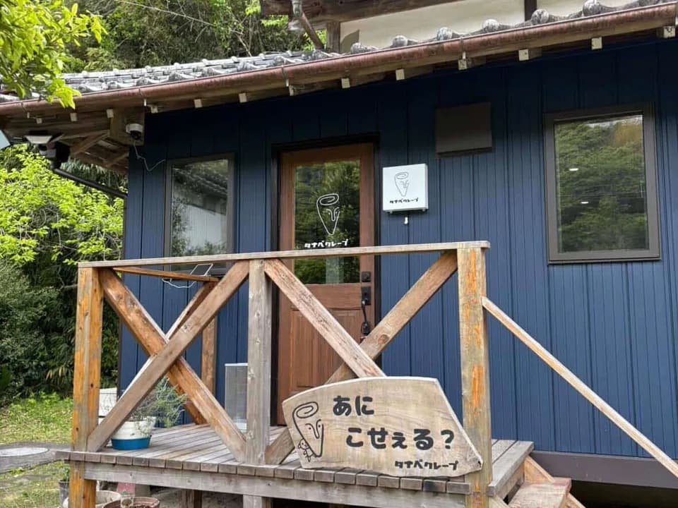 タナベクレープの店先。紺色の板壁に木の扉と窓、手前に木のデッキと手すり。デッキには房総の方言で「あに こせぇる?」と書かれた木の看板が立てかけてある