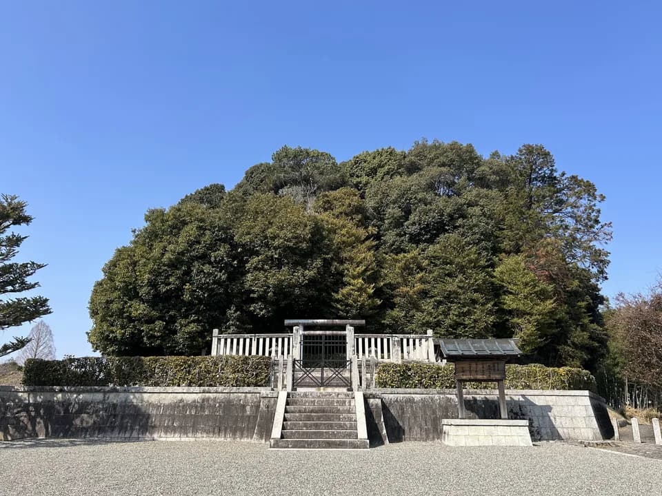 天武・持統天皇陵古墳の拝所を正面から見たところ。石段の上に白い柵と鉄の門扉があり、奥に濃い緑の木々に覆われた墳丘が盛り上がる。空は青く澄んでいる