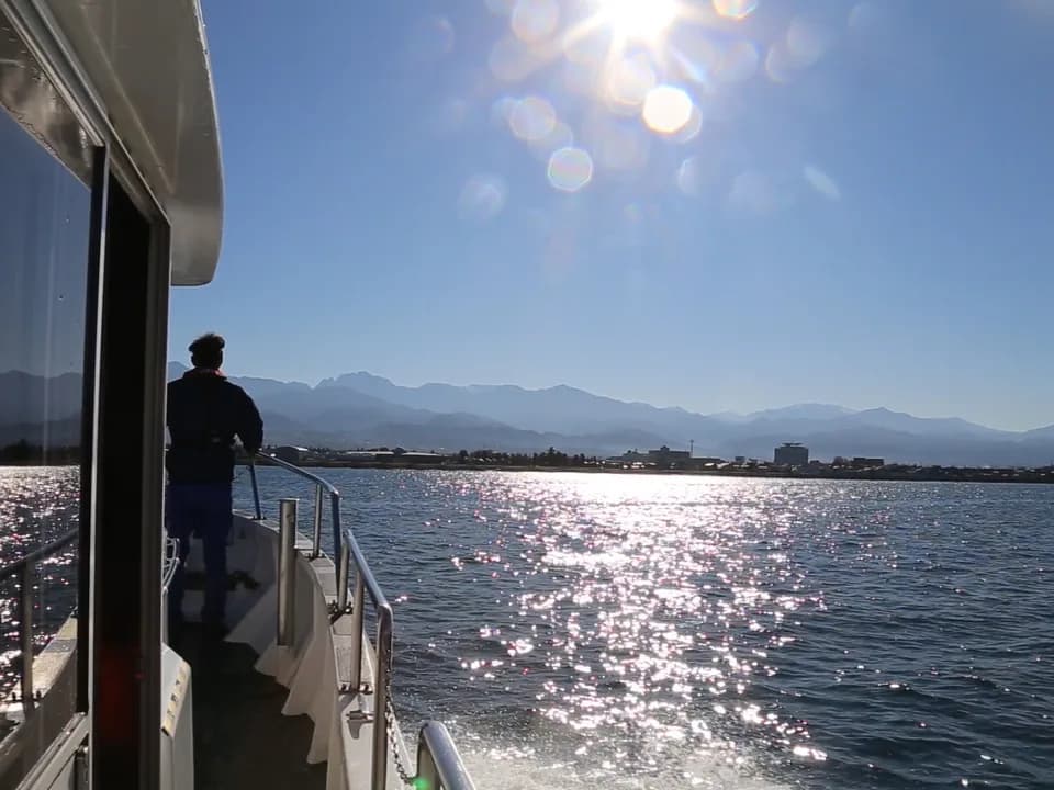 富山湾岸クルージングの船上から。逆光にきらめく海の先に立山連峰がかすみ、デッキに立つ乗客の後ろ姿