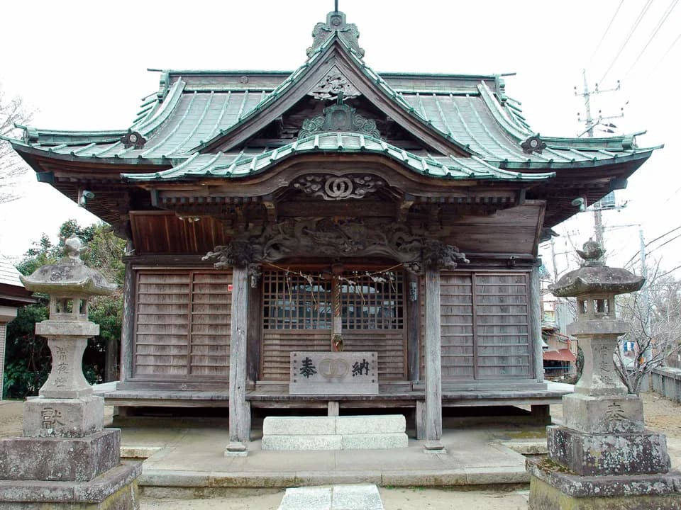 八坂神社の社殿を正面から見たところ。緑青色の瓦屋根を重ねた木造で、梁には龍の彫刻、前に一対の石灯籠が立つ