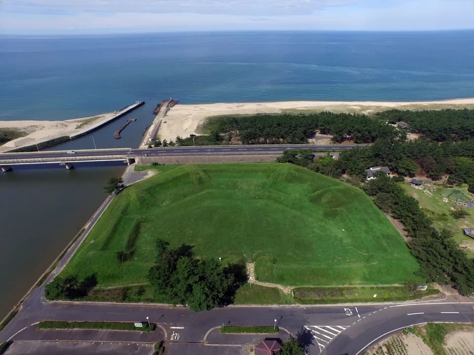 上空から見た由良台場。芝生に覆われた四角い土塁が広がり、その向こうに道路と松林、白い砂浜、青い日本海が続く